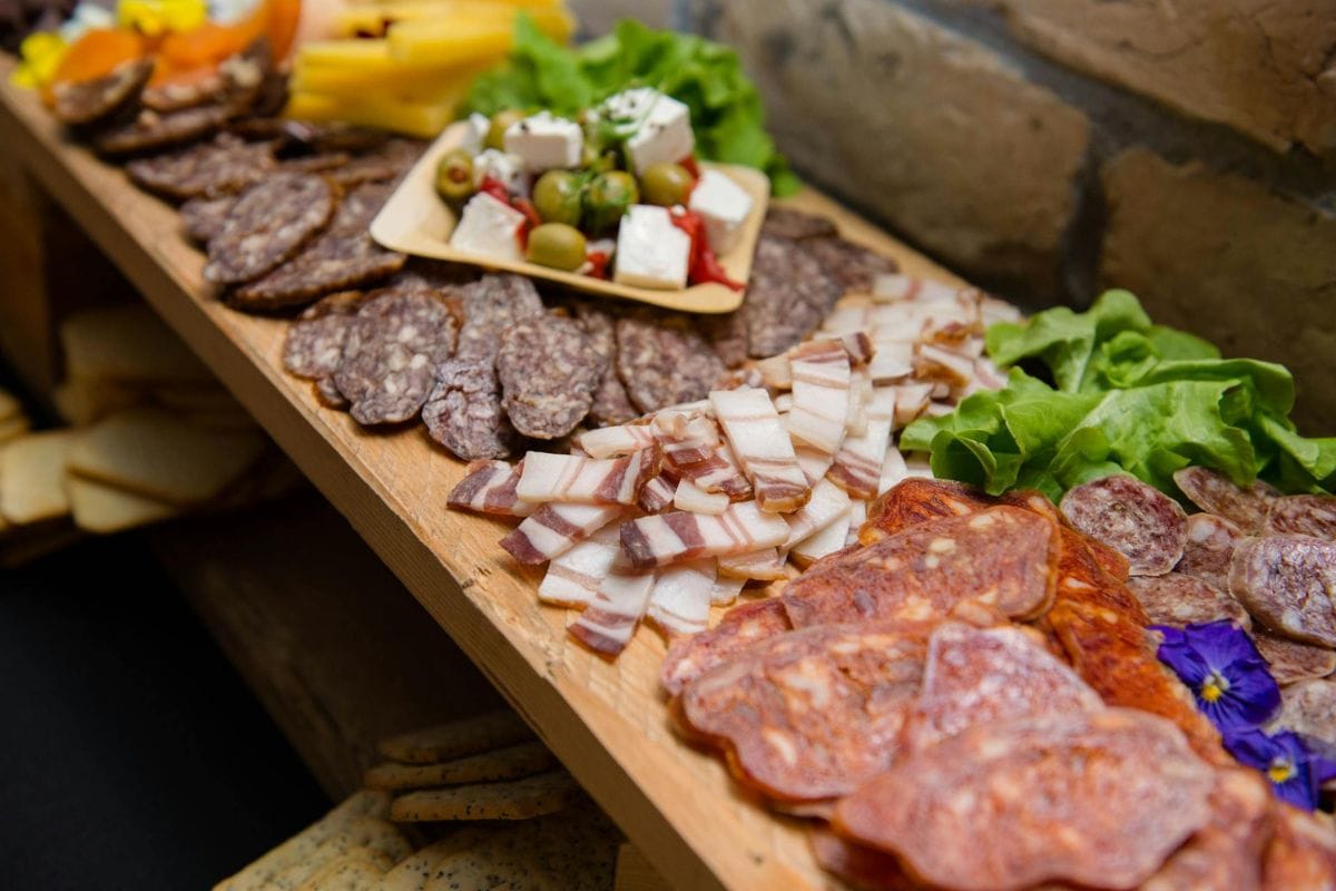 A wooden board with assorted cured meats, sliced on top, with a small dish of olives, feta, and vegetables, alongside lettuce and edible flowers.