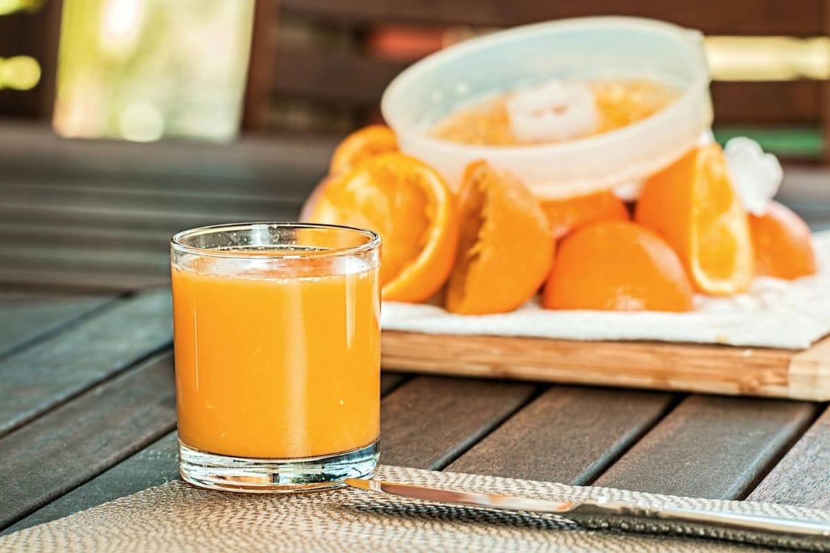 A glass of orange juice on a table with a knife. In the background, halved oranges and a juicer are placed on a wooden board.