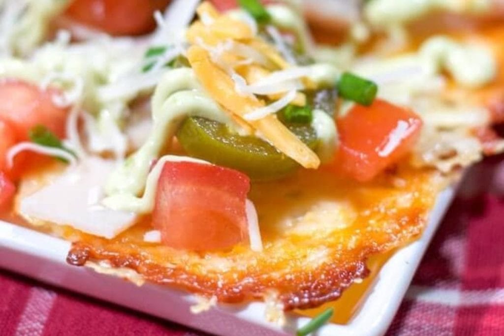 Close-up of a cheesy nacho topped with diced tomatoes, jalapeño slices, shredded cheese, and a drizzle of green sauce on a white dish.