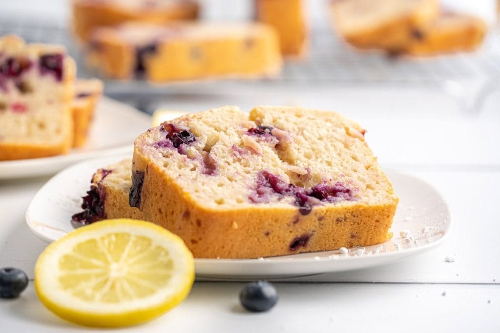 Slices of blueberry lemon bread on a white plate, with a lemon slice and scattered blueberries nearby.