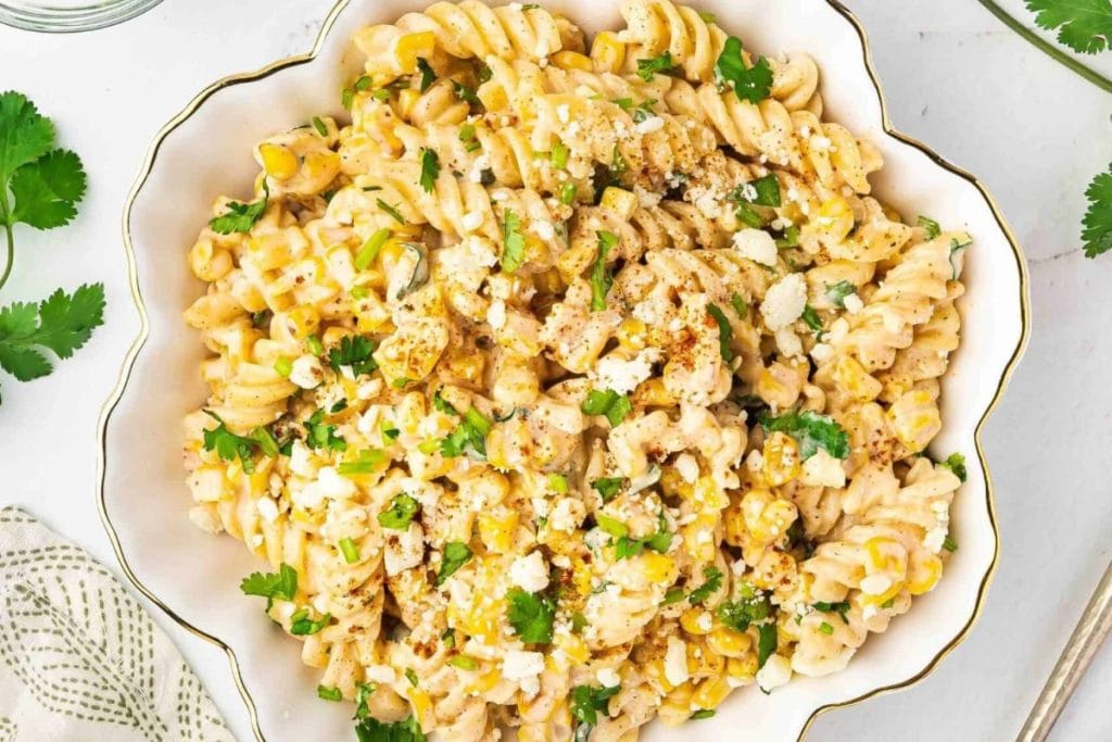 A bowl of pasta salad with fusilli, corn, crumbled cheese, and cilantro, garnished with pepper flakes.