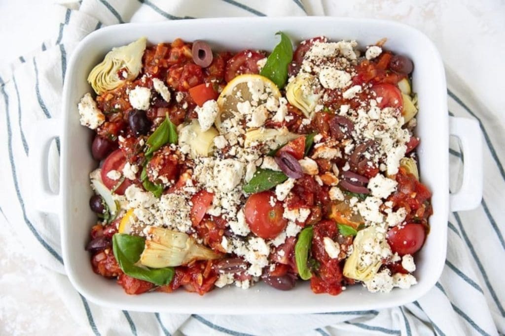Baked Mediterranean dish with artichokes, cherry tomatoes, olives, lemon slices, and crumbled feta cheese in a white dish on a striped cloth.