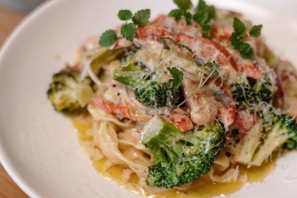 A plate of pasta topped with broccoli, sliced red peppers, and grated cheese, garnished with fresh greens.