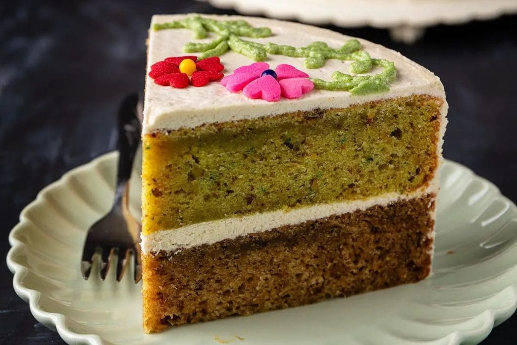 A slice of layered cake with white icing and colored flower decorations on top, placed on a white plate with a fork on the side.