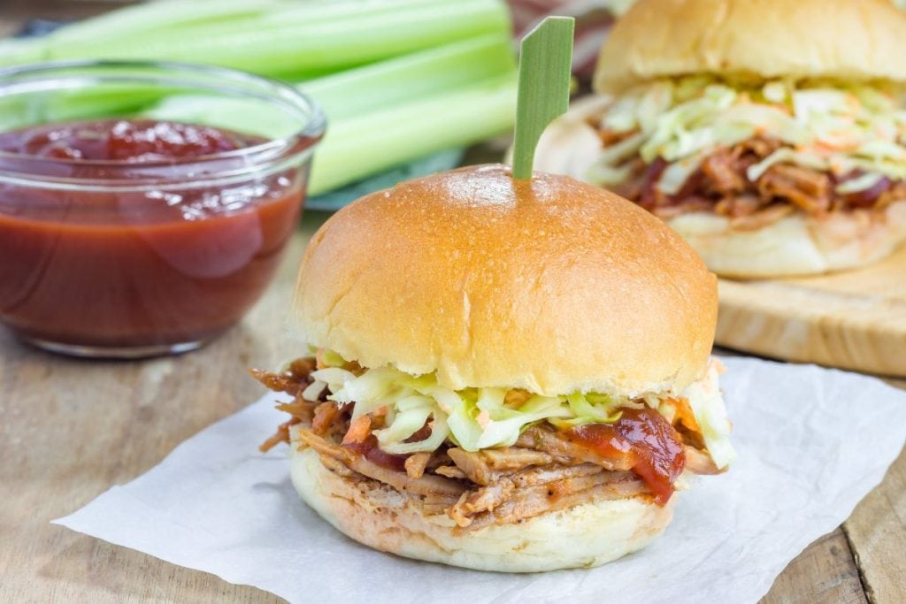 Pulled pork sandwich with coleslaw and BBQ sauce, garnished with a skewer, placed on parchment paper. A bowl of sauce and celery sticks are in the background.