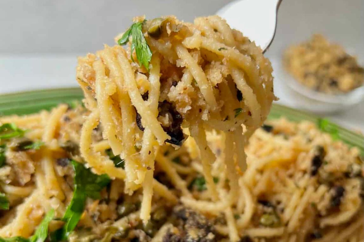 Forkful of spaghetti with breadcrumbs and herbs on a green plate, with a small bowl of breadcrumbs in the background.
