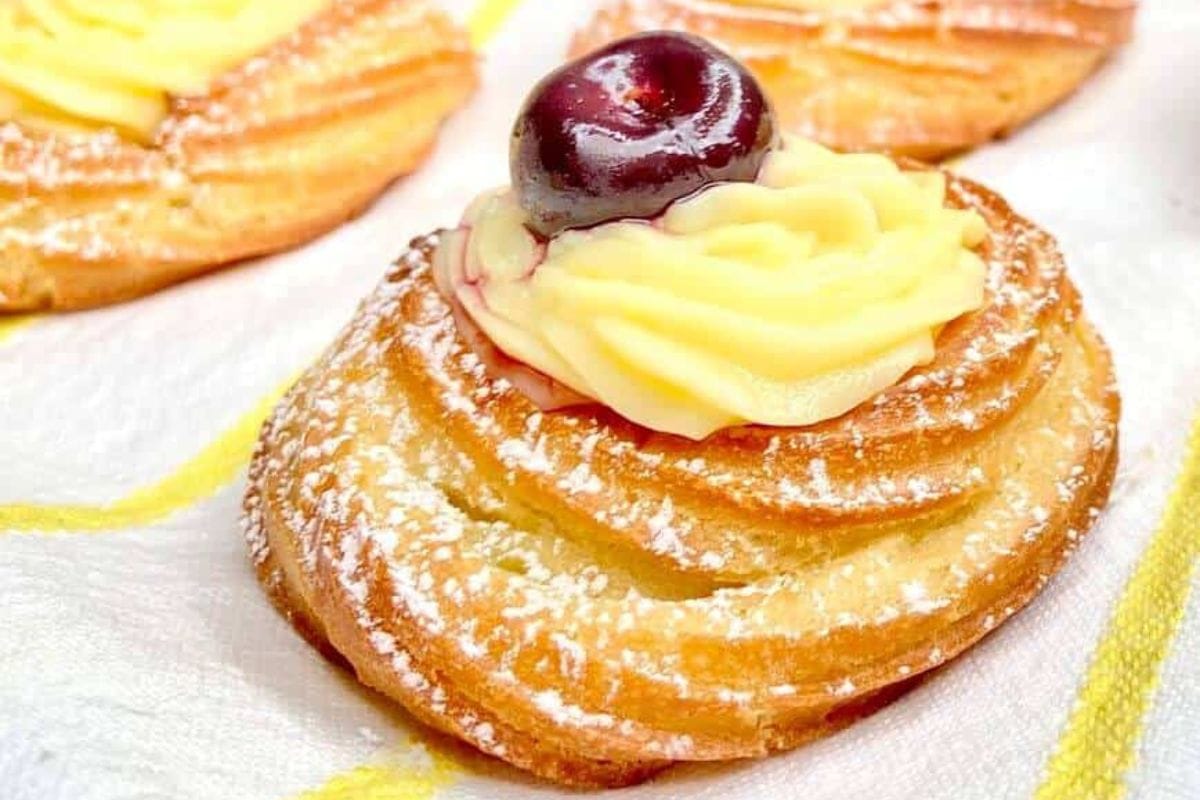 A pastry topped with a swirl of yellow cream and a dark cherry, dusted with powdered sugar, on a white surface.