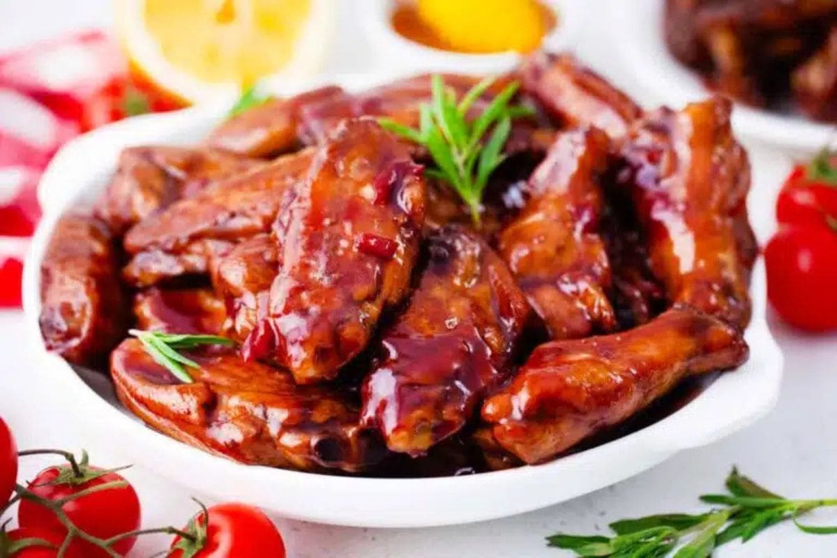 A white bowl filled with glazed chicken wings, perfect as appetizers for March Madness, is garnished with rosemary. Cherry tomatoes and lemon slices are visible in the background.