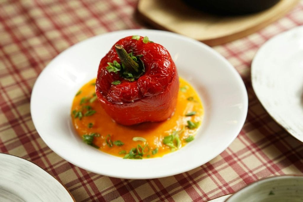 A red stuffed bell pepper sits on a white plate, surrounded by an orange sauce and garnished with chopped herbs, on a checkered tablecloth.