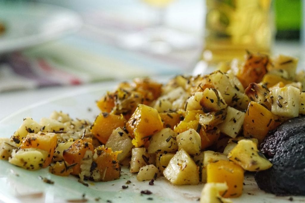 A plate with diced roasted vegetables, including squash and potatoes, seasoned with herbs, next to a slice of grilled meat.