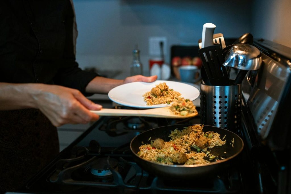 Person serving rice and vegetable dish from a pan onto a white plate in a kitchen setting.