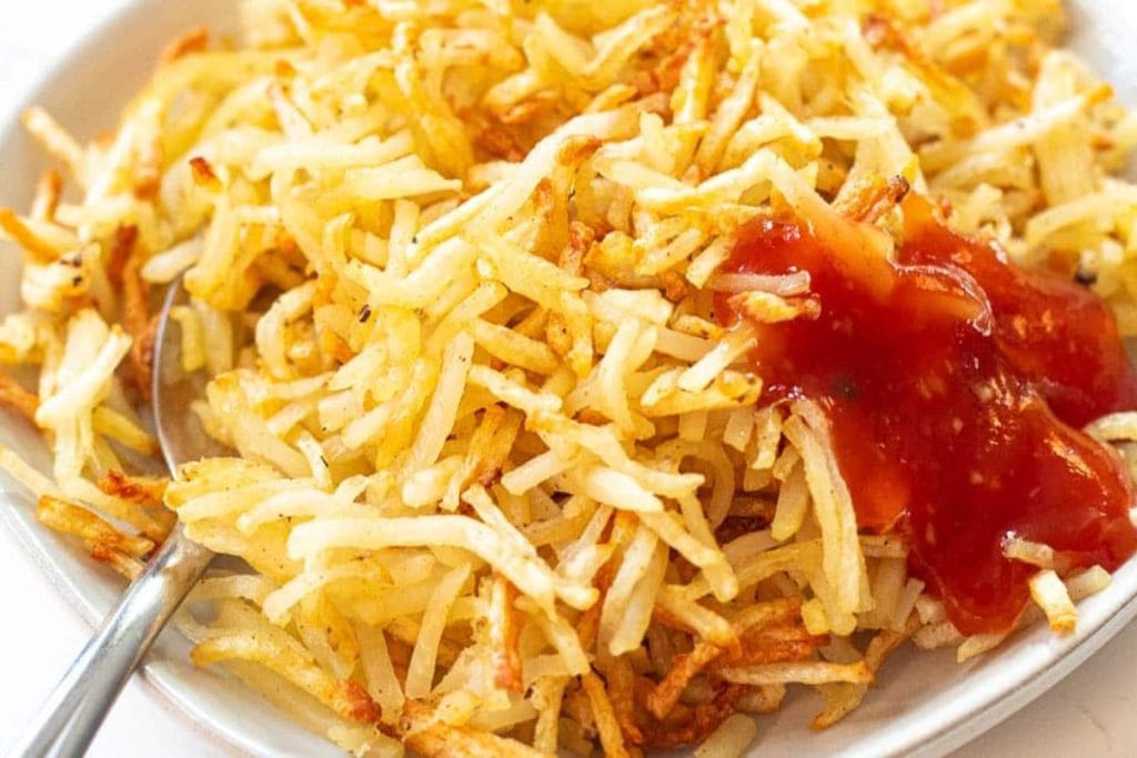 A plate of golden brown hash browns with a spoon, accompanied by a serving of ketchup on the side.