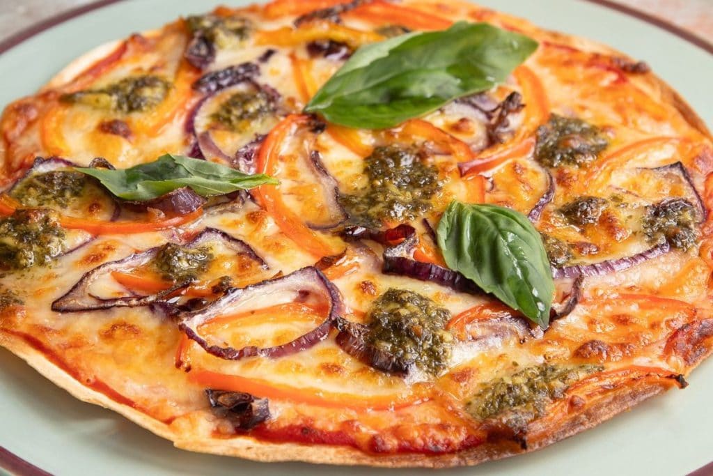 A thin-crust pizza topped with red onions, red bell peppers, pesto, melted cheese, and fresh basil leaves, served on a green plate.