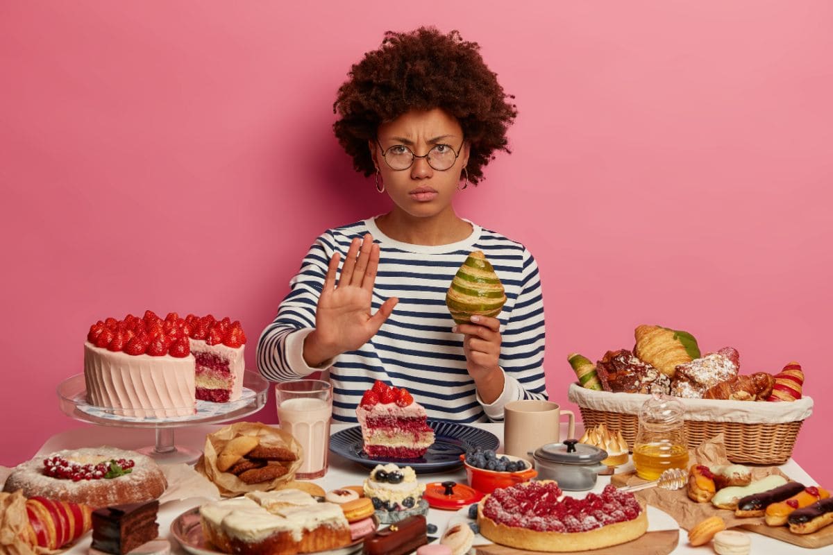 A person wearing glasses and a striped shirt holds up a hand, signaling stop, while sitting at a table full of various desserts against a pink background.
