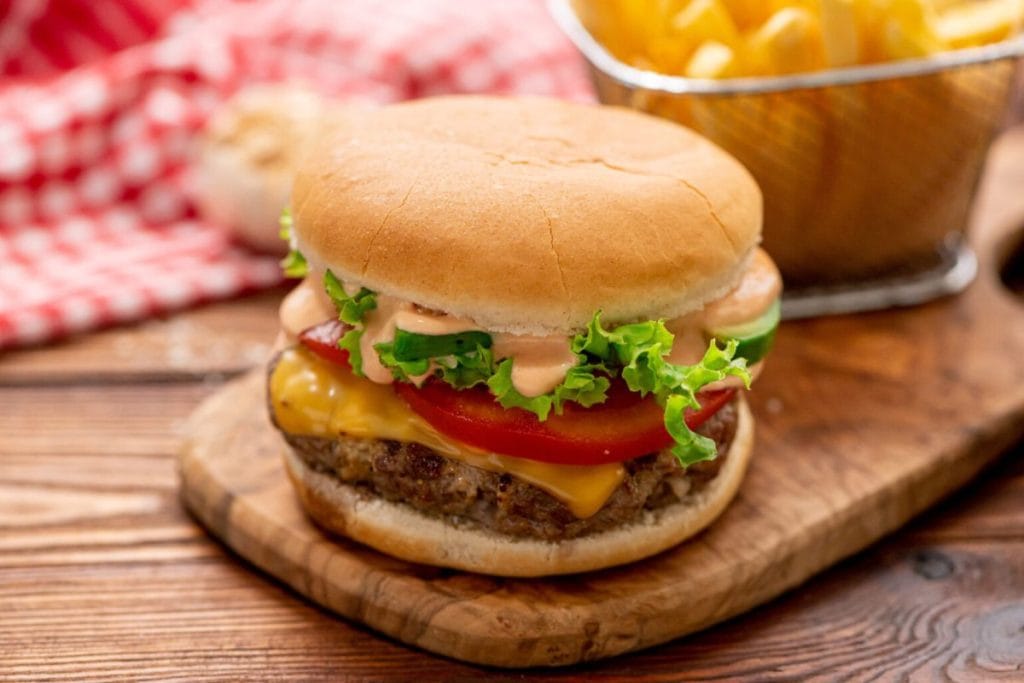 A cheeseburger with lettuce, tomato, and sauce on a wooden board, next to a basket of fries and a red checkered cloth.