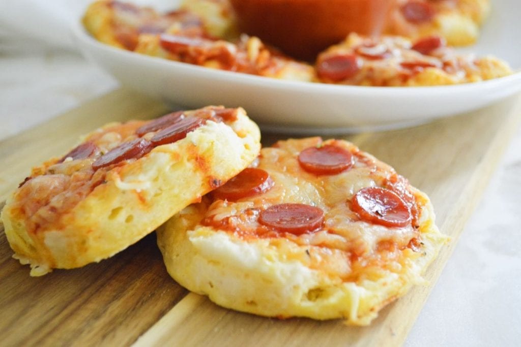 Two mini pepperoni pizzas are placed on a wooden board, with more pizzas on a white plate in the background.
