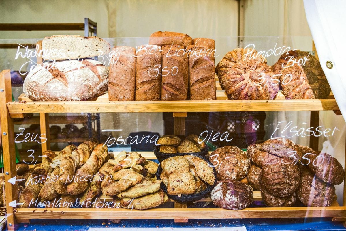 Breads displayed on wooden shelves in a bakery. Each type has a chalk-written label and price on the glass, including pumpkin bread, rye bread, and various other artisanal loaves.