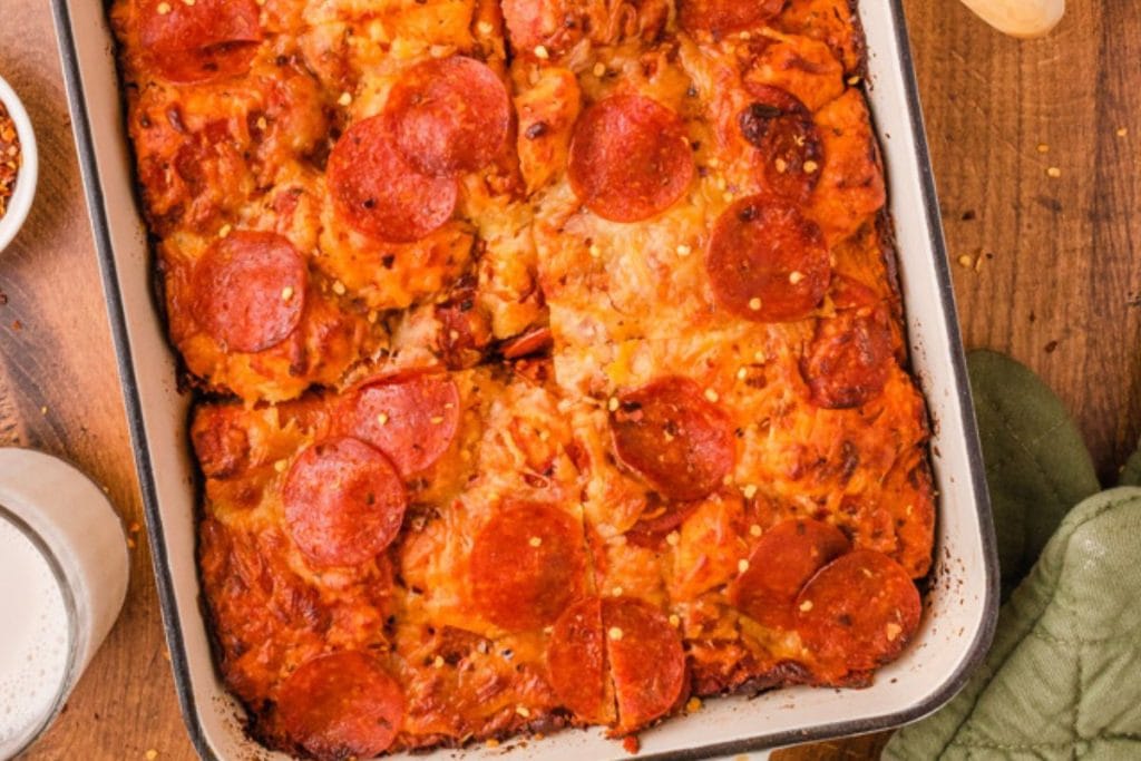 Rectangular pepperoni pizza in a baking dish on a wooden surface, with a small bowl of red pepper flakes nearby.