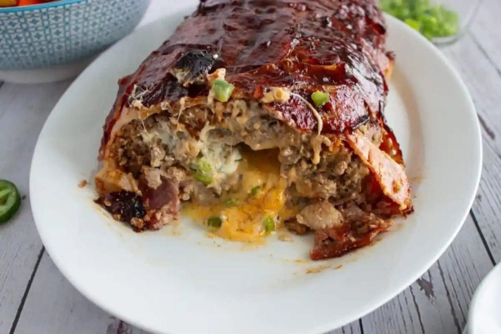 Sliced bacon-wrapped meatloaf filled with cheese and green peppers on a white plate, with a bowl in the background.