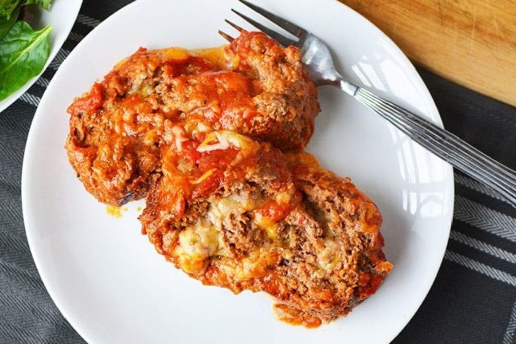 Two slices of cheesy, saucy meatloaf on a white plate with a fork, placed on a dark tablecloth.