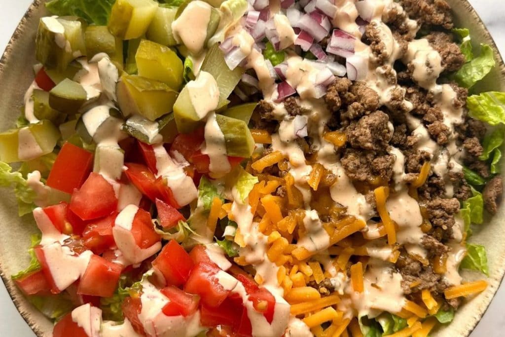 A salad with lettuce, diced tomatoes, cheese, ground beef, chopped pickles, and onions, topped with a creamy dressing.