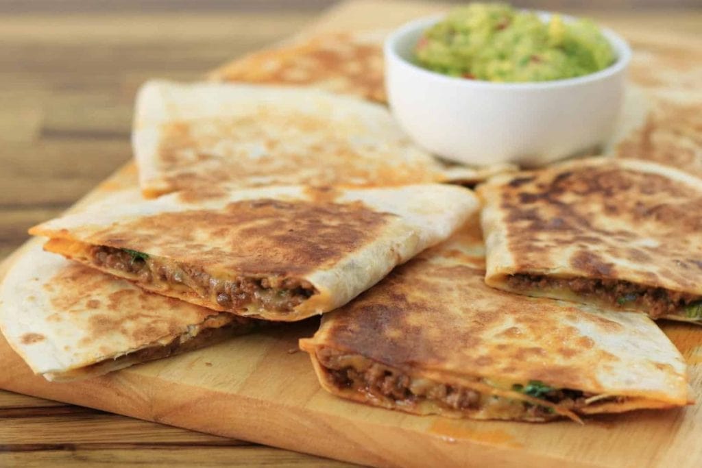 Sliced quesadillas with a meat and cheese filling are arranged on a wooden board. A small bowl of guacamole is placed in the background.