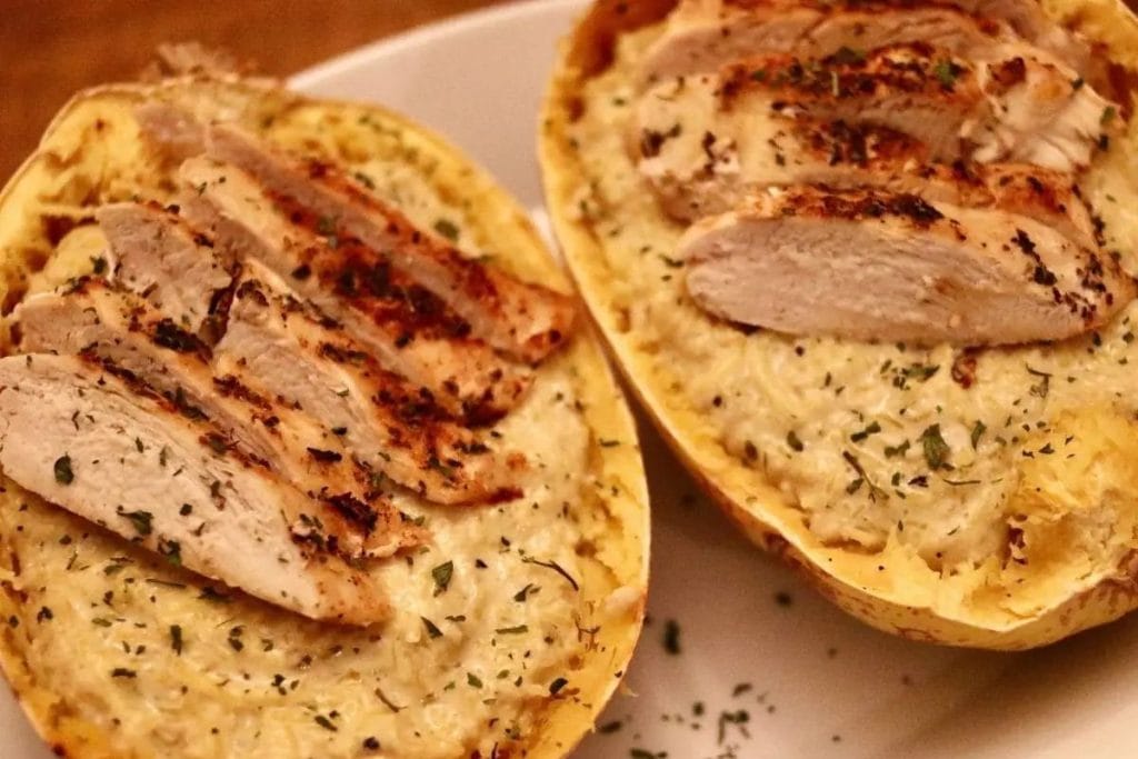 Two halved spaghetti squash filled with creamy pasta and topped with sliced grilled chicken, garnished with herbs on a white plate.