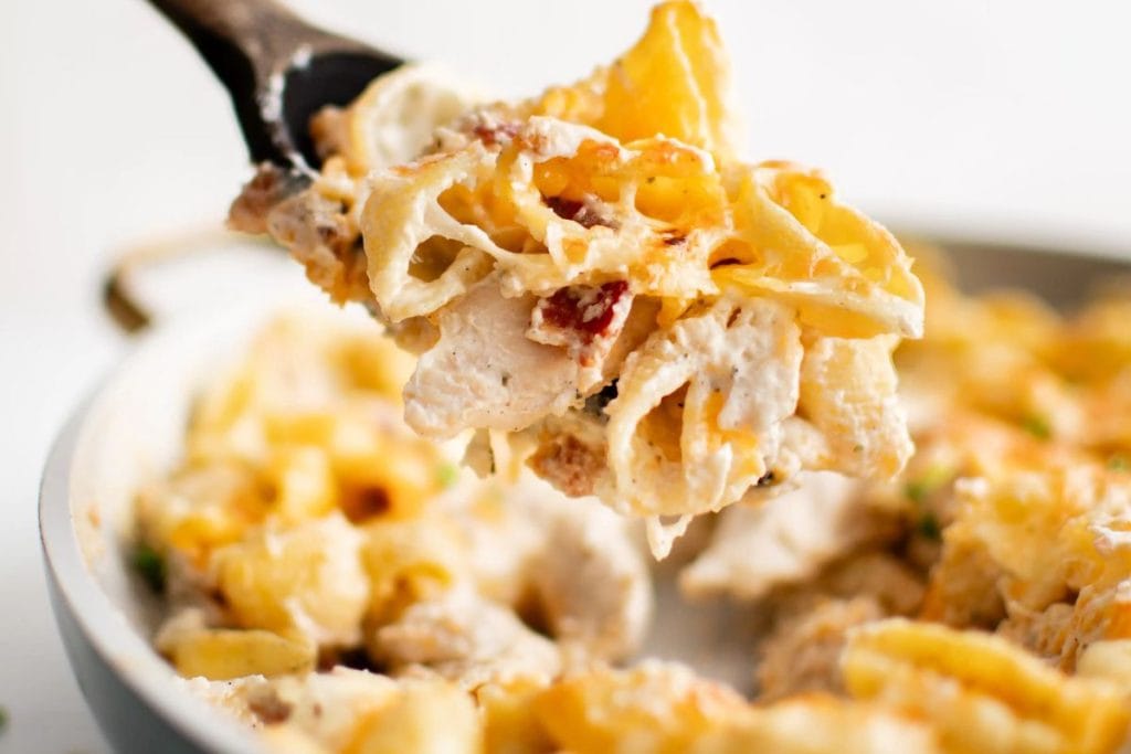 Close-up of a serving of creamy chicken pasta with bacon bits being scooped from a skillet with a wooden spoon.