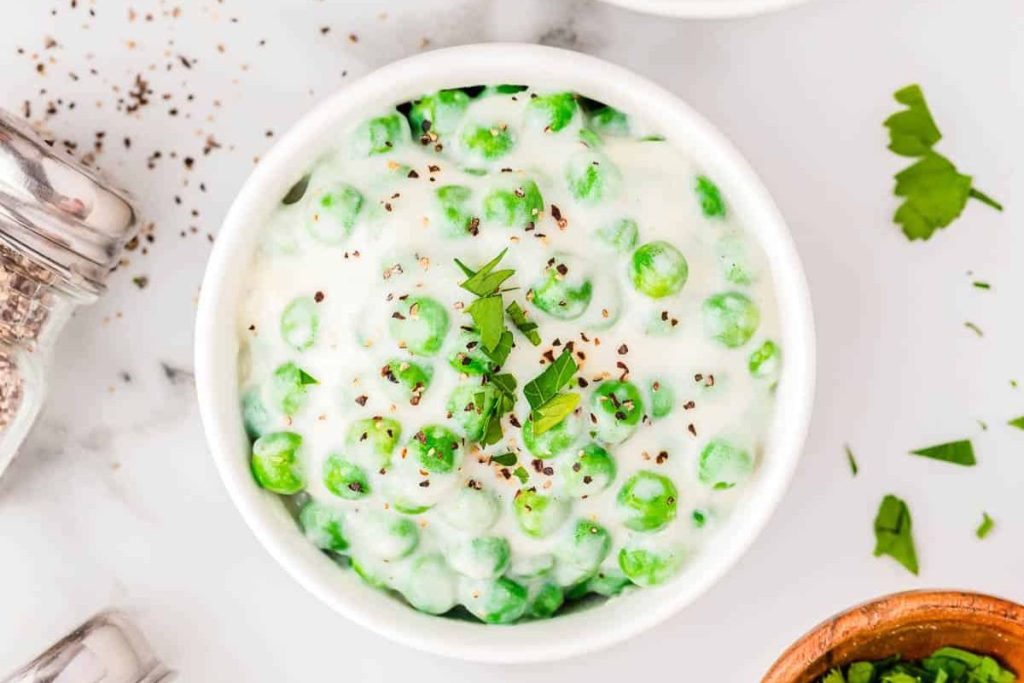 Bowl of peas in a creamy white sauce, garnished with chopped parsley and black pepper, on a marble surface.