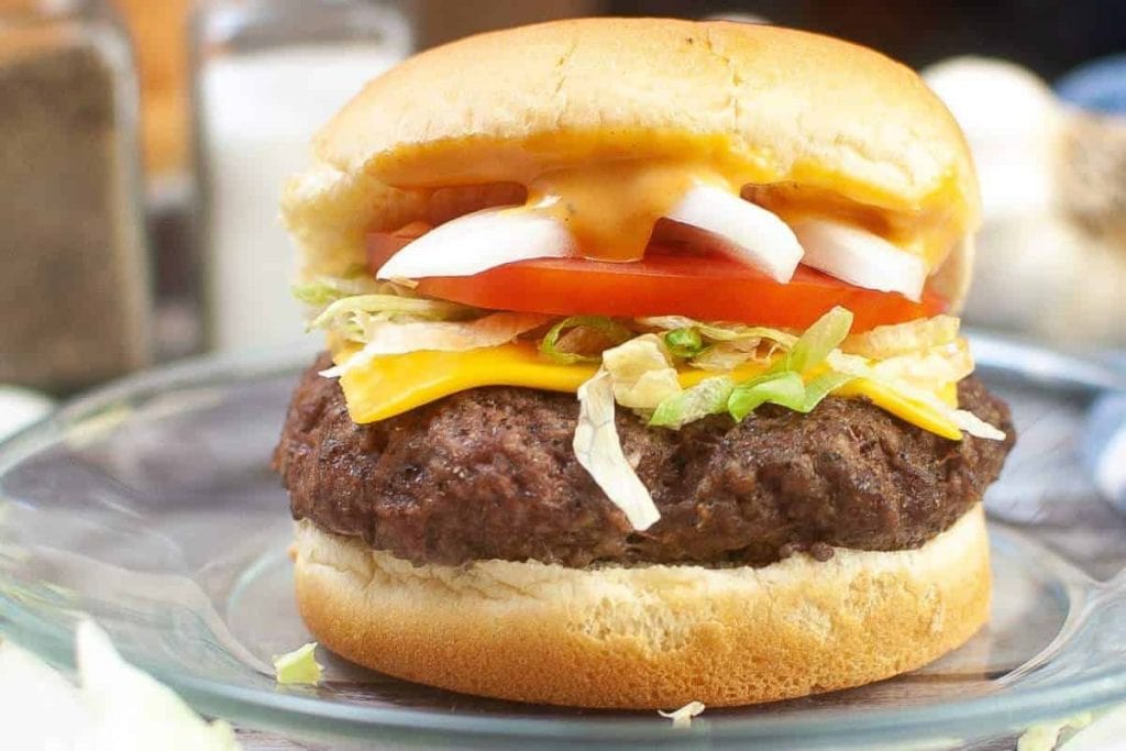 A cheeseburger with lettuce, tomato, onions, and sauce, placed on a clear glass plate.