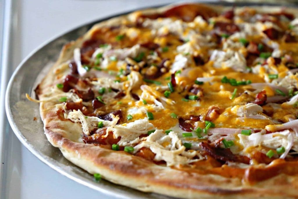 Close-up of a pizza with shredded chicken, chunks of bacon, melted cheese, and chopped green onions on a metal tray.