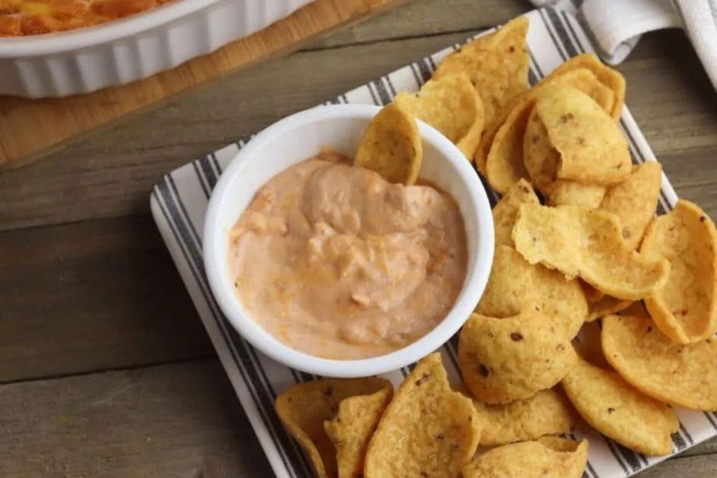 A bowl of creamy dip surrounded by scoop-shaped corn chips on a striped towel on a wooden surface.