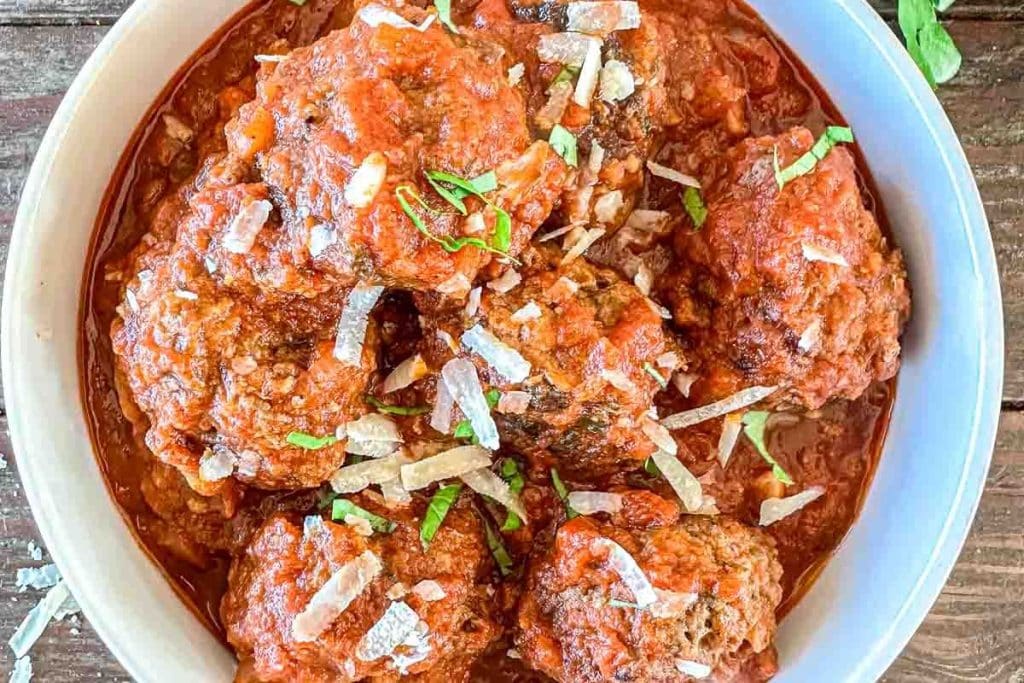 A bowl of meatballs covered in tomato sauce, garnished with shaved cheese and chopped herbs, placed on a wooden surface.