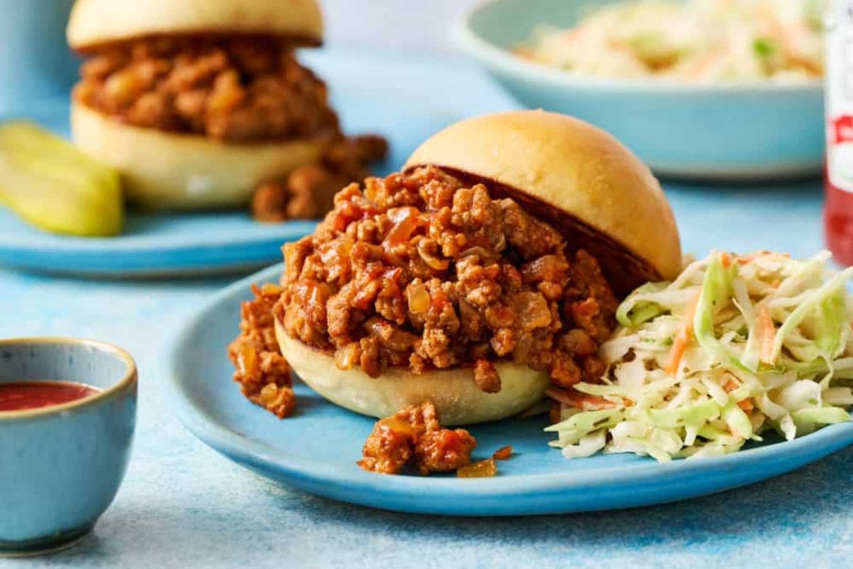A sloppy joe sandwich on a blue plate with coleslaw on the side. A pickle and another sandwich in the background.