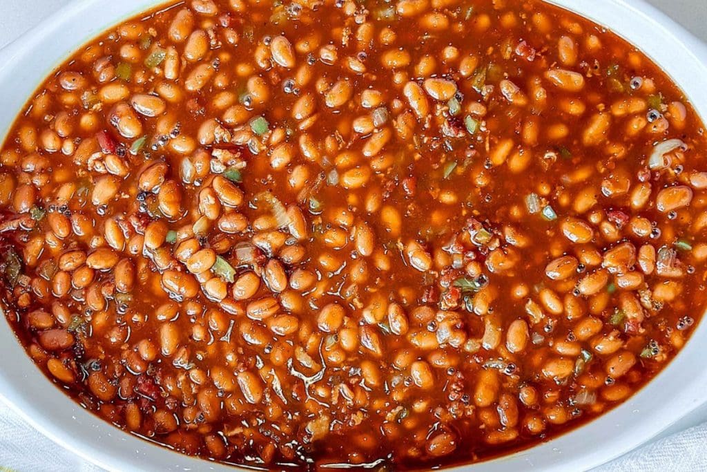 A close-up of baked beans in a white dish, featuring a rich tomato-based sauce and small pepper pieces.