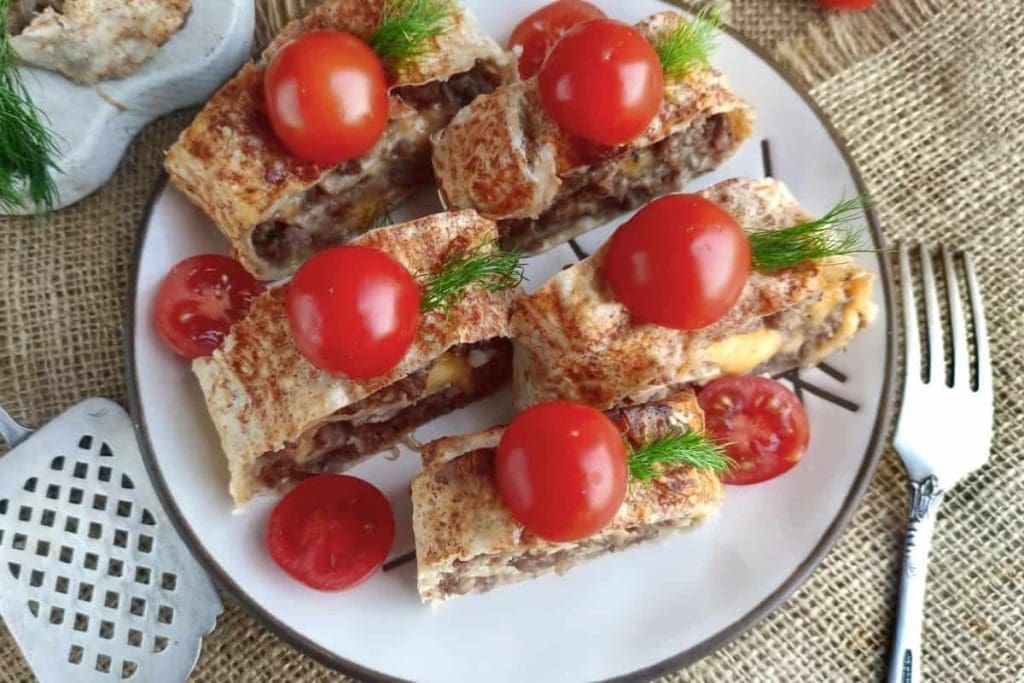 Slices of savory pastry filled with meat, topped with cherry tomatoes and herbs, served on a white plate.