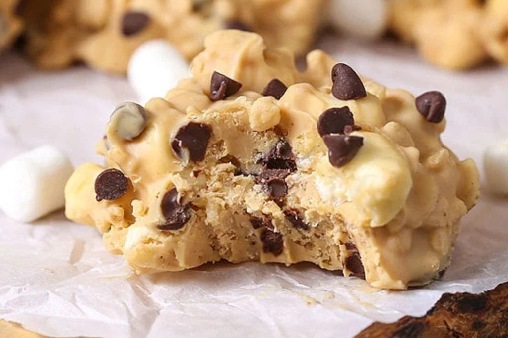 A cookie with chocolate chips and a bite taken out, surrounded by mini marshmallows on parchment paper.