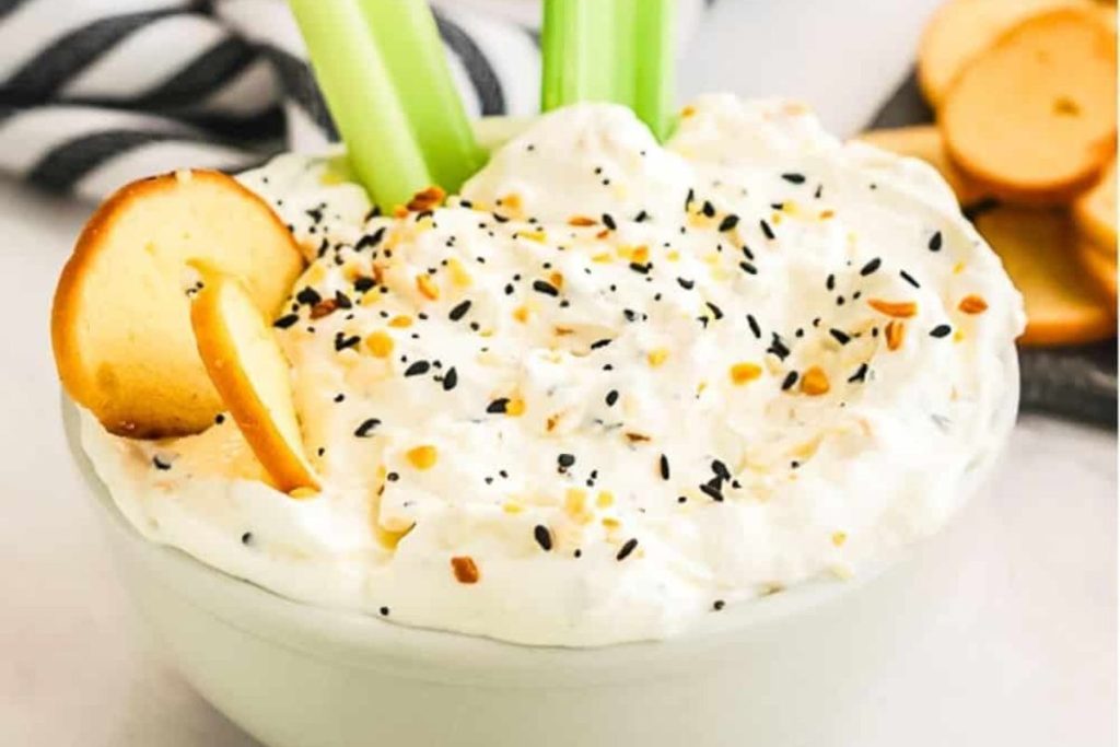 Creamy dip in a bowl topped with everything bagel seasoning, a bagel chip, and celery sticks; assorted bagel chips on the side.