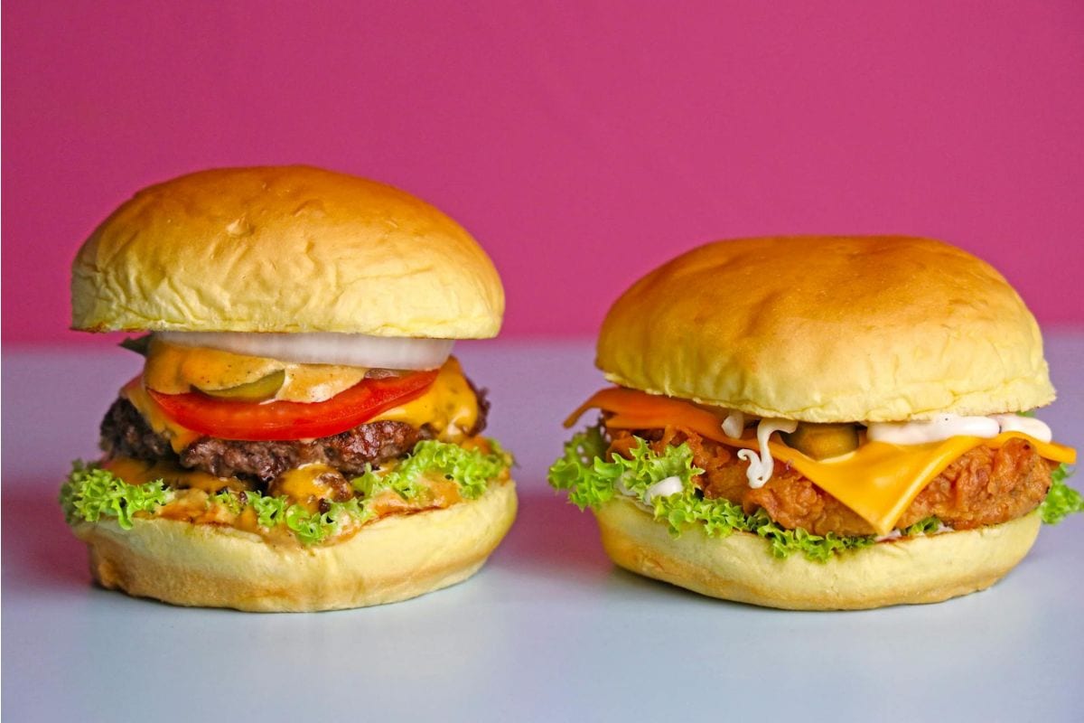 Two burgers on a light surface with a pink background: one beef burger with lettuce, tomato, onion, and cheese; one chicken burger with lettuce, cheese, and sauce.