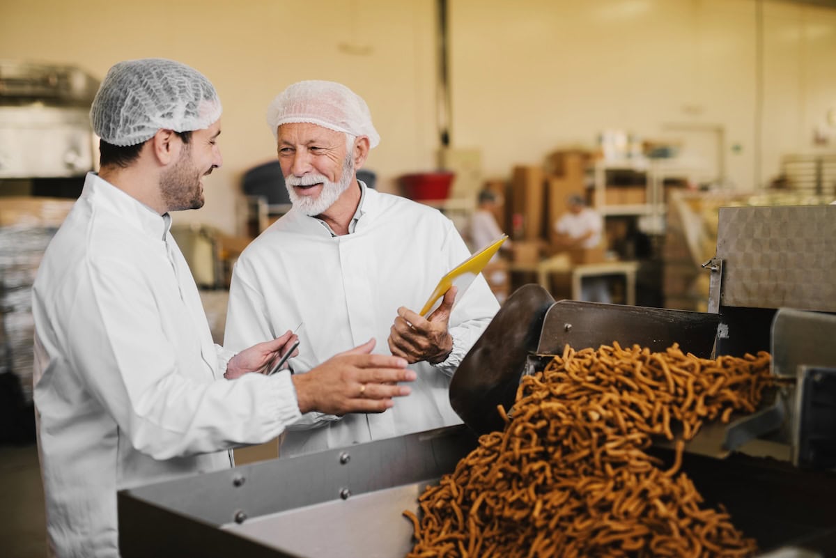 Two men in white coats and hairnets converse in a factory, standing next to a machine producing twisted snacks. One man holds a clipboard.