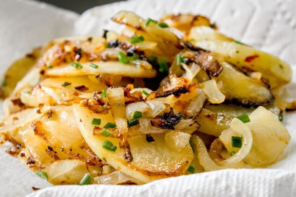 A close-up of cooked potato slices with browned onions and chopped chives on top, resting on a paper towel.