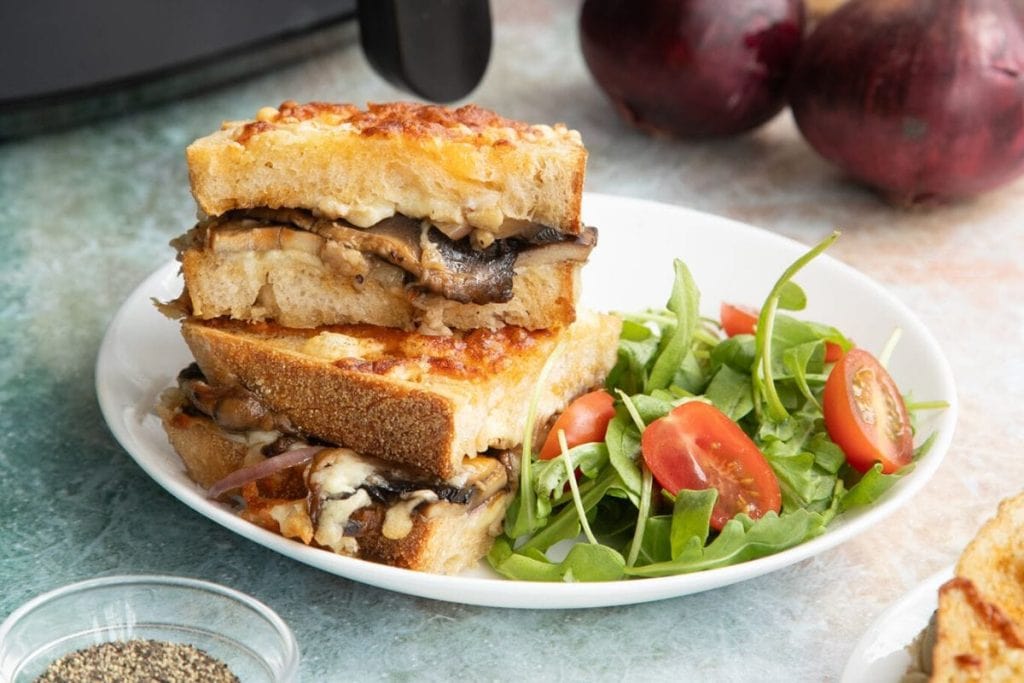 Grilled cheese sandwich with mushrooms on a white plate, served with a side salad of arugula and cherry tomatoes. Red onions and a small bowl of pepper are in the background.