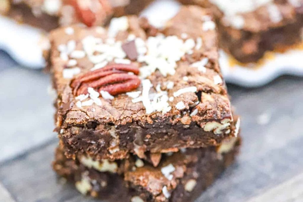 Close-up of two chocolate brownies stacked, topped with pecans and shredded coconut, on a wooden surface. More brownies are blurred in the background.