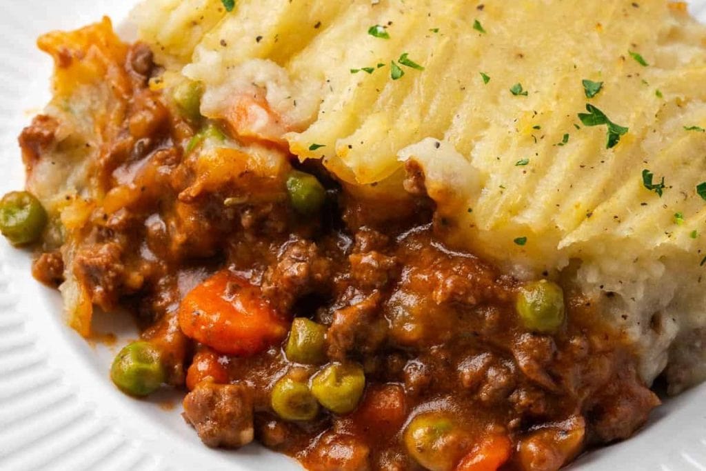 A serving of shepherd's pie on a white plate, featuring mashed potatoes, ground beef, peas, and carrots, garnished with chopped parsley.