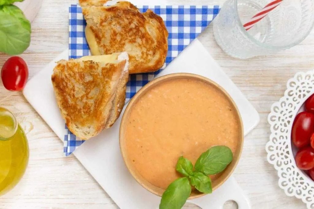 Bowl of tomato soup garnished with basil next to a grilled cheese sandwich on a blue checkered napkin, with cherry tomatoes and a glass of water in the background.