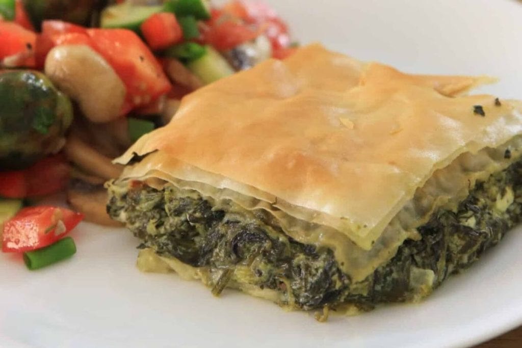 Slice of spinach pie with flaky pastry on a plate, accompanied by a vegetable medley including tomatoes, mushrooms, and green peppers.