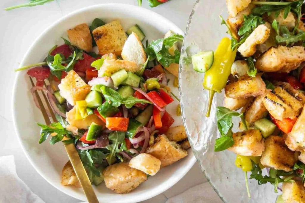 A bowl of salad with grilled bread, arugula, cucumber, tomatoes, red onions, and bell peppers, next to a large serving bowl with more salad.