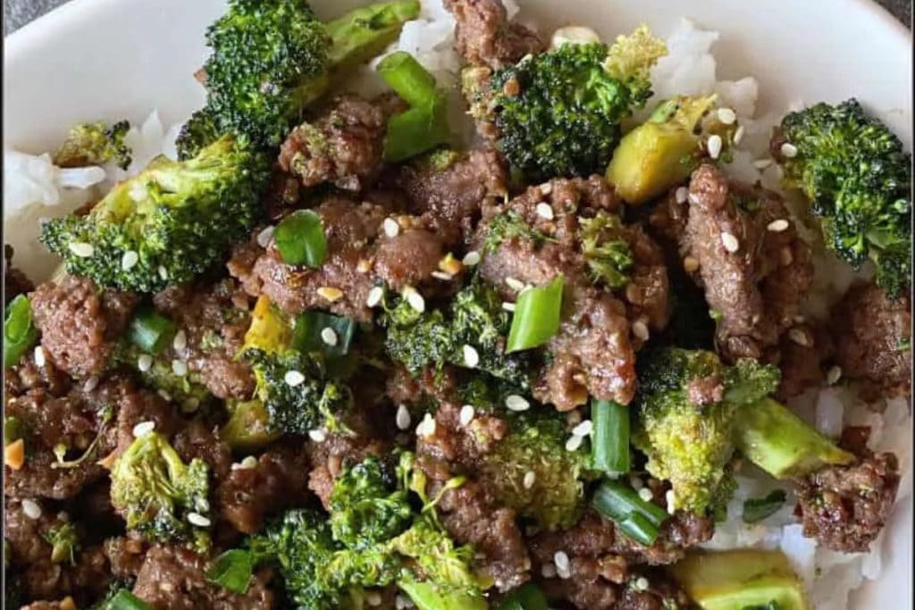 A bowl of white rice topped with beef and broccoli, garnished with green onions and sesame seeds.