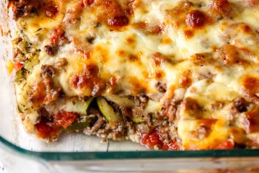 A close-up of a baked lasagna in a glass dish, showing layers of melted cheese, ground meat, and tomato sauce. A portion is missing from the corner.