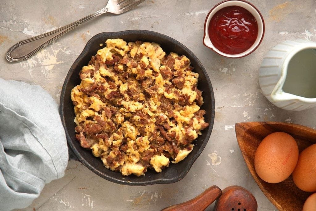 Skillet with scrambled eggs and sausage next to ketchup, pitcher, silver fork, cloth, whole eggs, and wooden salt shaker on a textured surface.